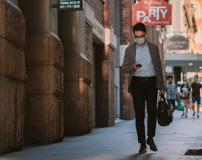Man wearing face mask walking down a modern alley way with a brief case whilst on his phone