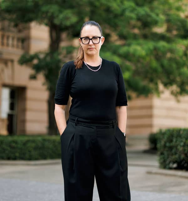 A woman named Danielle in black pants and glasses standing in front of a building.