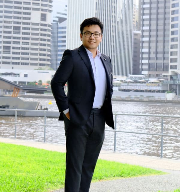 A man in a business suit standing by a river with city buildings in the background, photographed by Neil Yang.