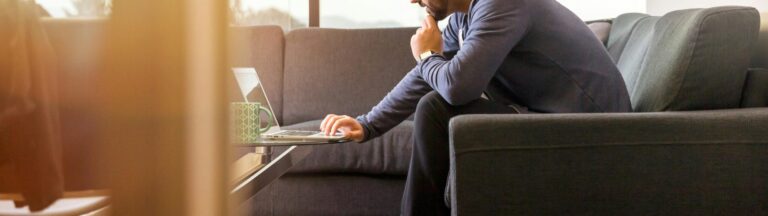 A man in a blue hoodie sits on a gray couch, working on a laptop placed on a glass coffee table in a well-lit room, planning the restructure of his small business in Australia.