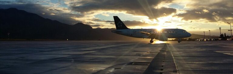 An airplane is parked on a tarmac at sunset in Australia, with the sun partially hidden behind the aircraft and mountains visible in the background.